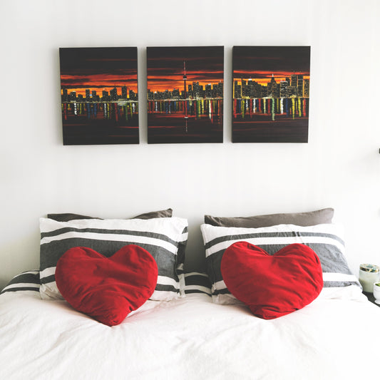 Bedroom with heart-shaped red pillows on a bed, nightstands with books and lamps, and wall art of a cityscape.